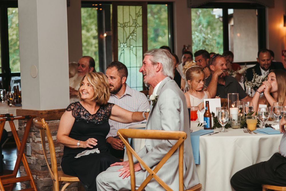 Wedding reception guests seated at long tables in an indoor venue with natural lighting and festive atmosphere.
