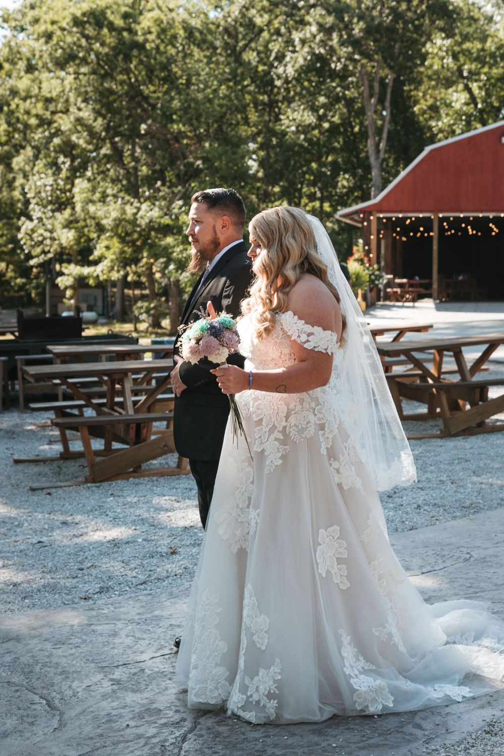 Couple stands together holding bouquet at rustic outdoor venue with picnic tables.
