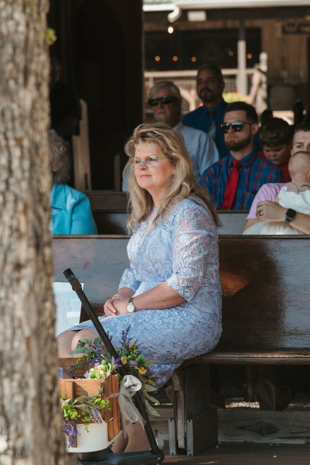 Guest in lace dress seated on wooden bench at outdoor wedding venue.