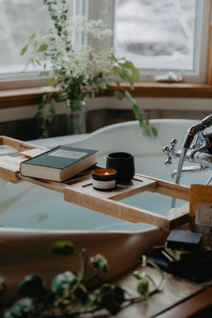 Cozy bath caddy with book, candle and fresh flowers by window with natural light.
