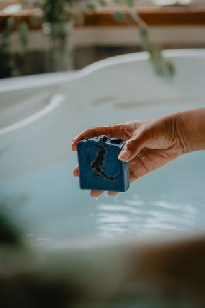 Hand holding a blue bath bomb over water with soft natural lighting.