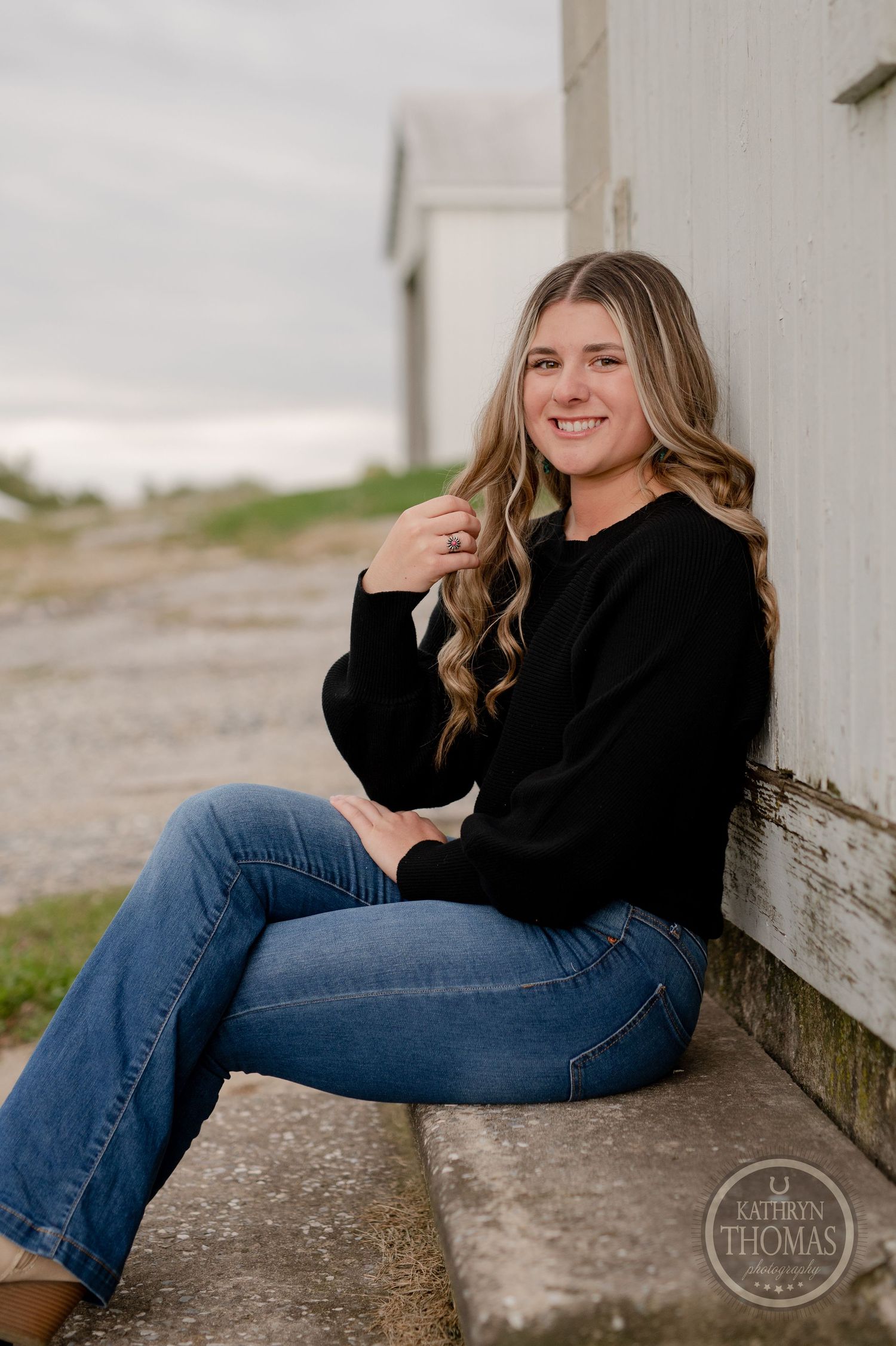 Outdoor portrait featuring a casual look with black sweater and jeans while sitting against a white wall background.