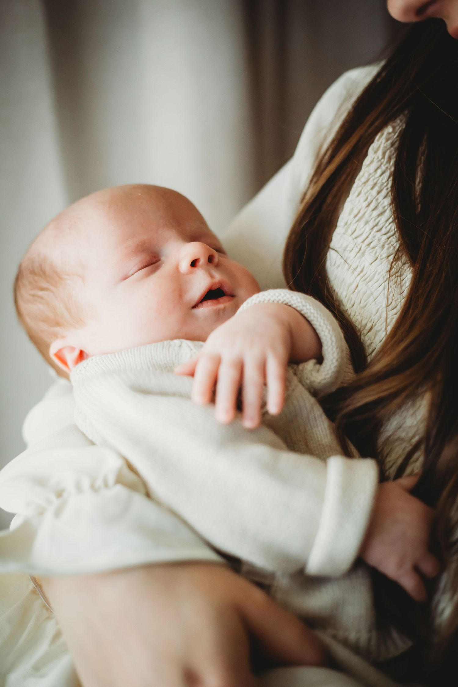 Peaceful newborn sleeps in white outfit against caregiver's cream sweater.