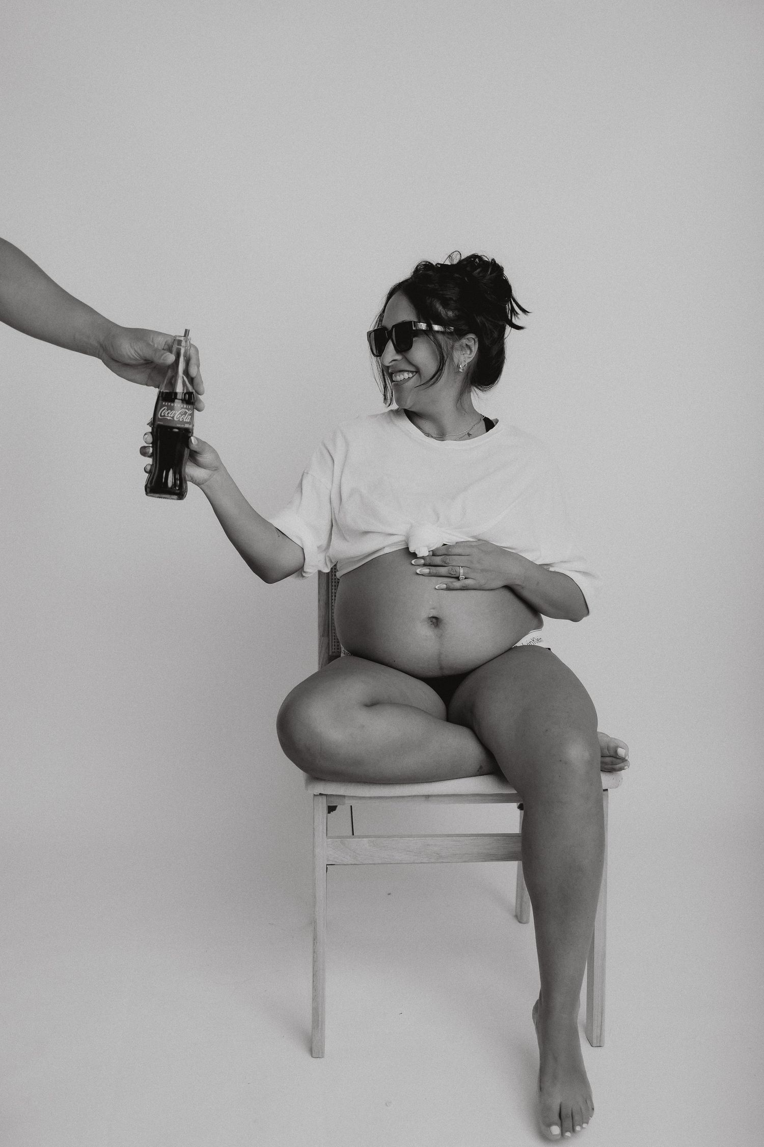 Casual maternity photo of expectant mother in white shirt accepting beverage while seated.