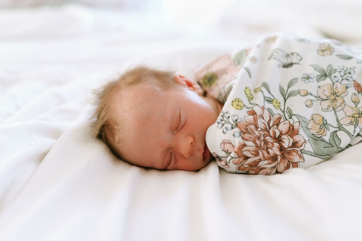 Peaceful newborn sleeps swaddled in floral print blanket on white bedding.