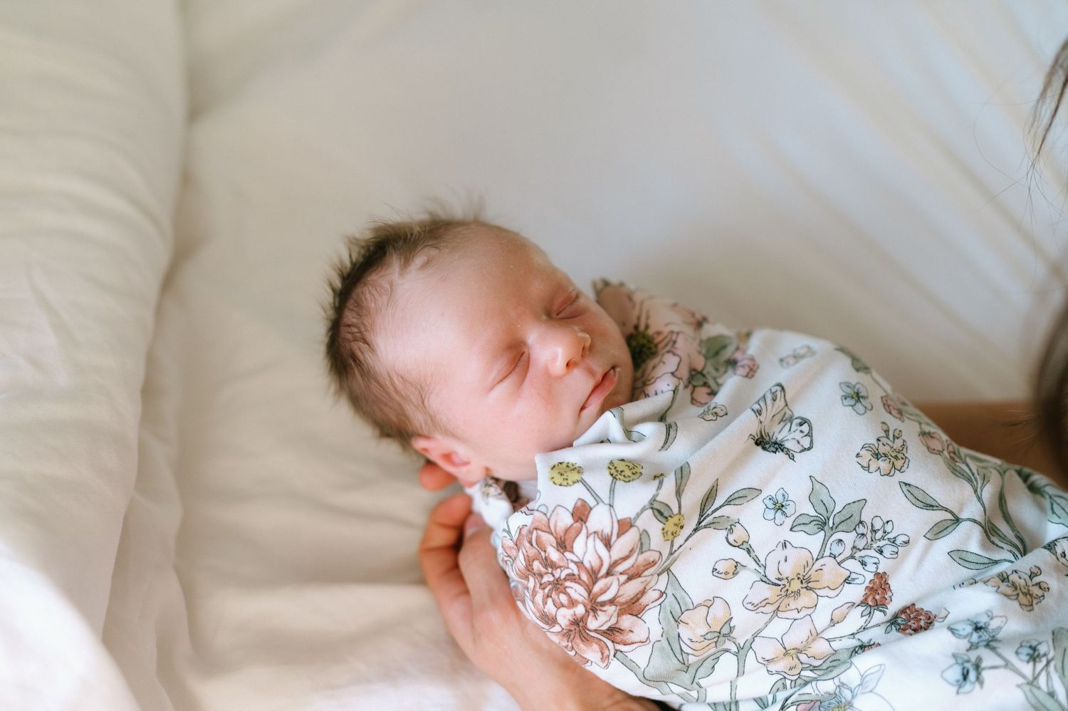 Sleeping baby wrapped in delicate floral swaddle on white sheets.
