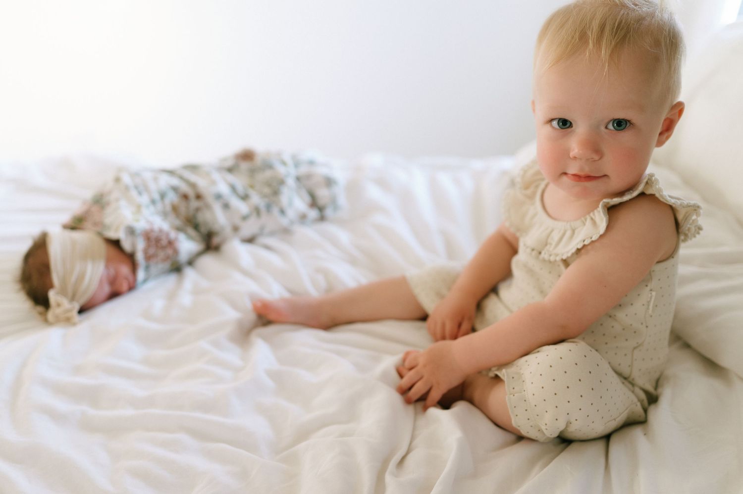 Toddler wearing cream-colored outfit sits upright on white bedding.