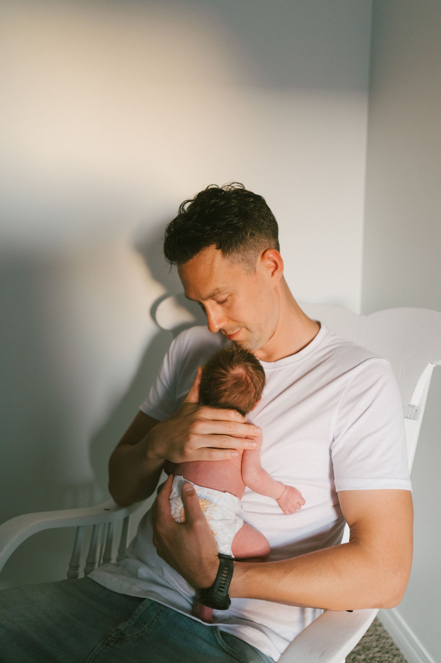 Parent tenderly holding sleeping newborn baby in white rocking chair by window.