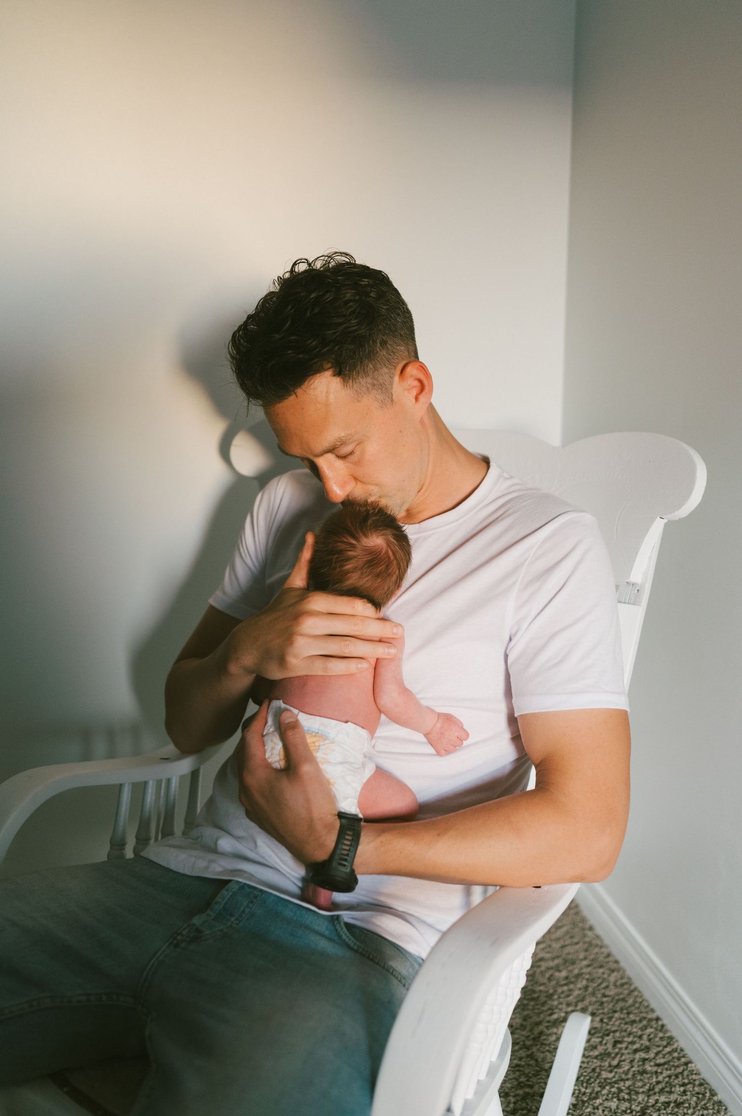 Parent cradling newborn infant while sitting in white chair near window with soft lighting.