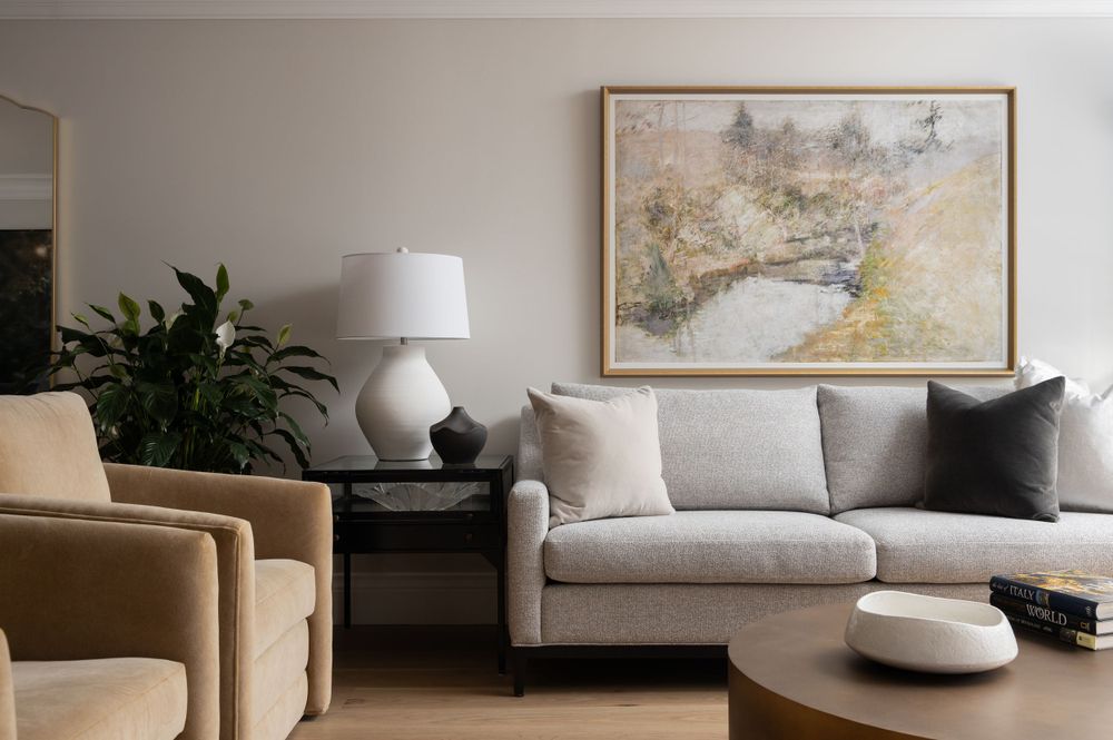 Modern living room with gray sofa, accent chair, and abstract landscape artwork on beige wall. Moody interior design photography. Toronto.