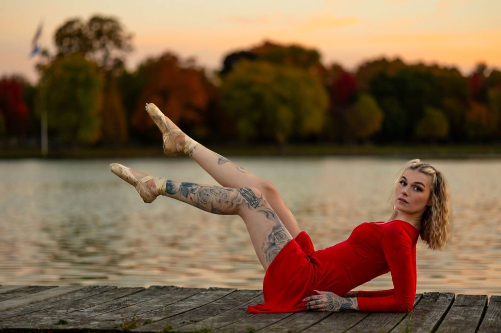 Ballet dancer in red dress performs graceful pose on wooden dock at sunset by lake with autumn trees.
