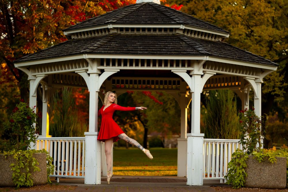 Ballerina in red dress strikes elegant pose in white gazebo surrounded by fall foliage.