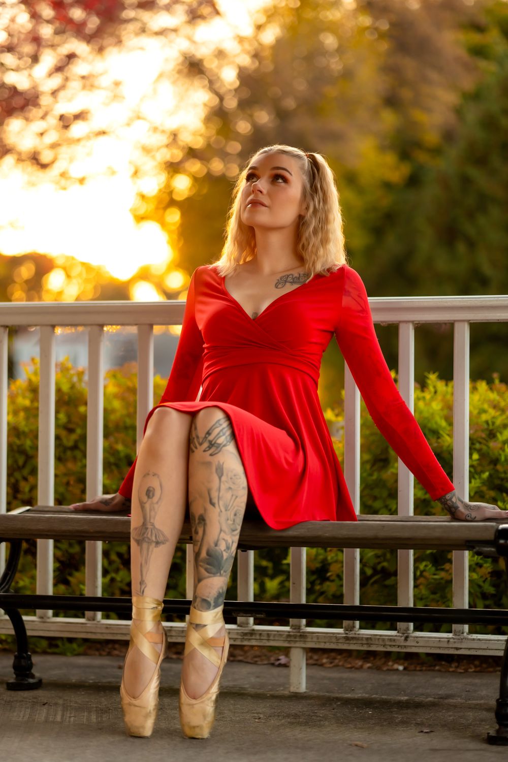 Dancer in red dress sits on railing wearing pointe shoes against golden sunset backdrop.