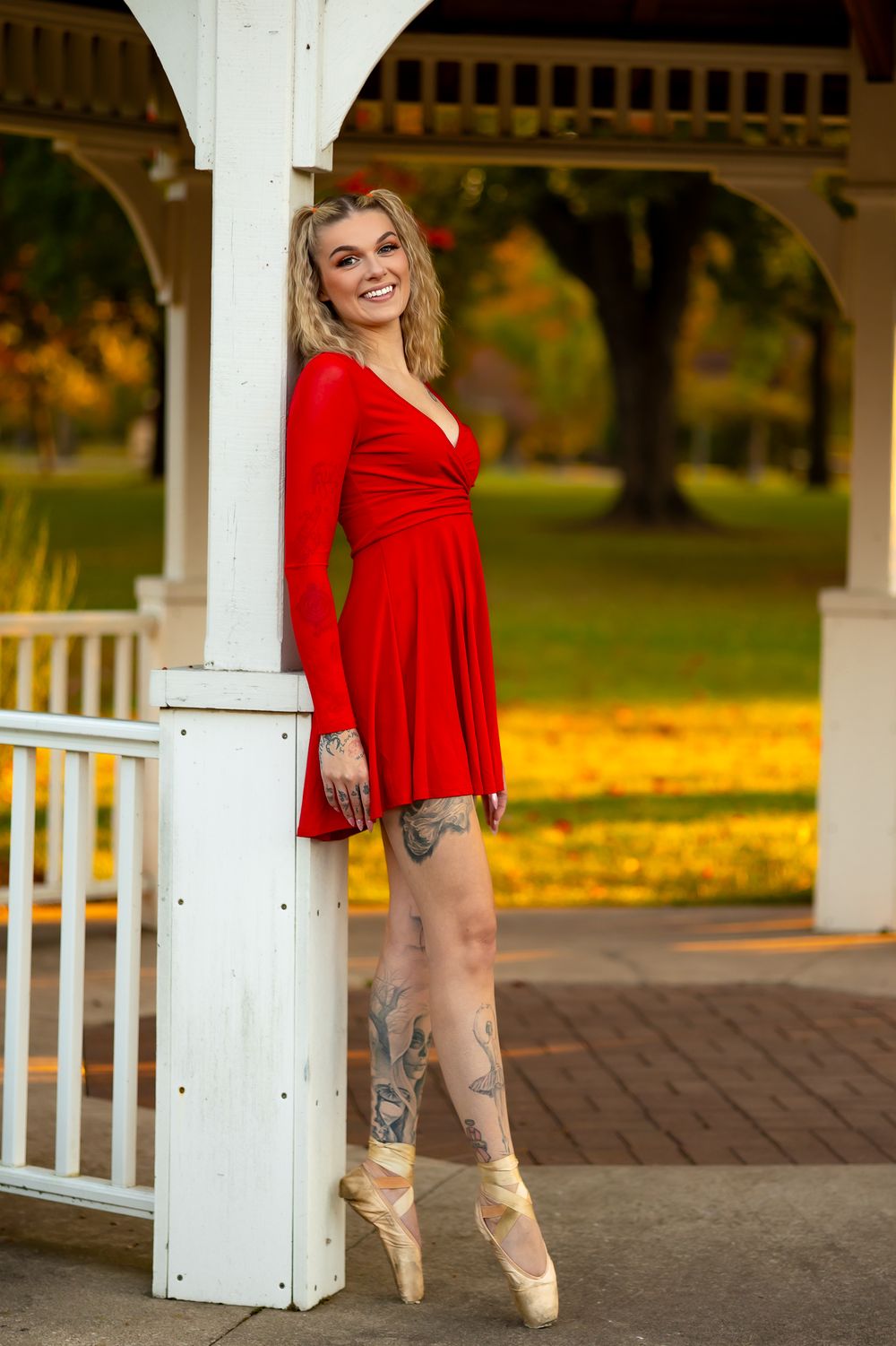 Ballerina poses en pointe in red dress against white gazebo column during autumn.