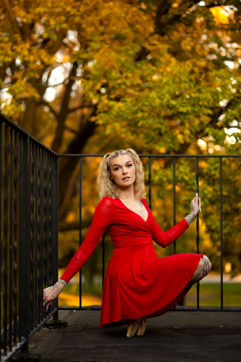 Ballet performer in red dress strikes artistic pose against iron railing with autumn trees behind.