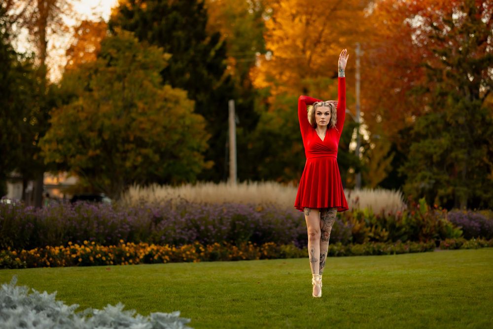 Ballerina performs graceful leap in red dress against autumn garden backdrop.