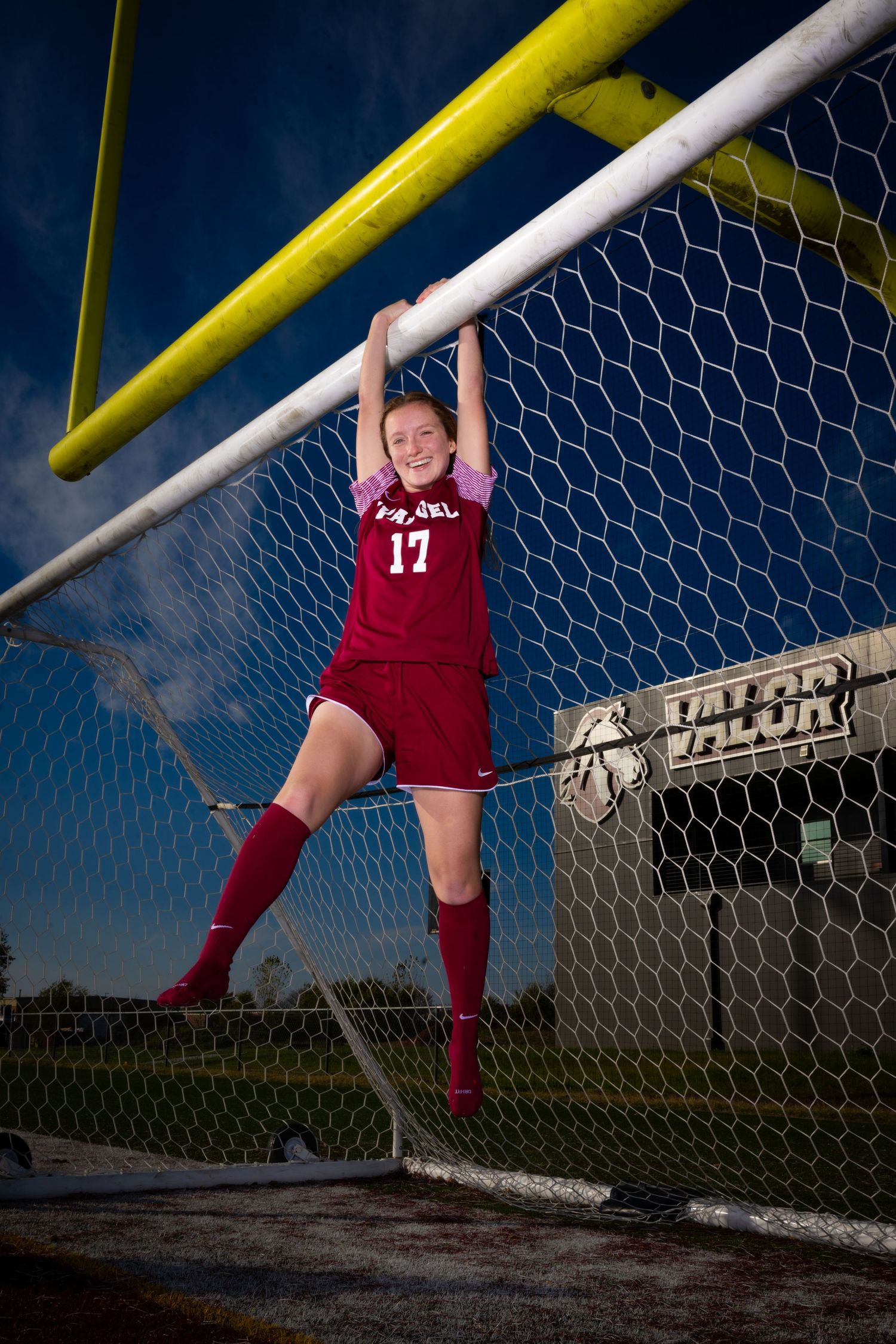 A soccer player in red uniform hangs from the goal post crossbar against a blue sky.
