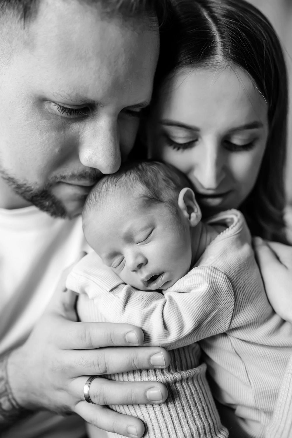 Black and white intimate portrait of parents cradling their sleeping newborn baby in a tender embrace.
