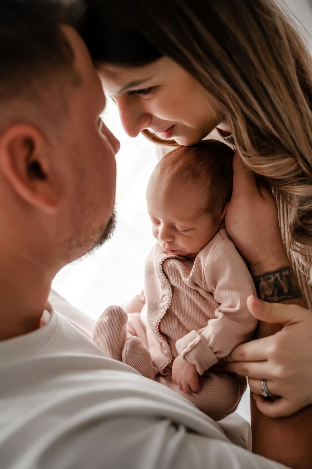 A tender family moment as parents lovingly gaze at their sleeping newborn baby in a soft pink outfit.