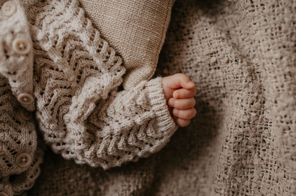 A tiny baby hand reaches out through the soft folds of a cozy knitted beige sweater.