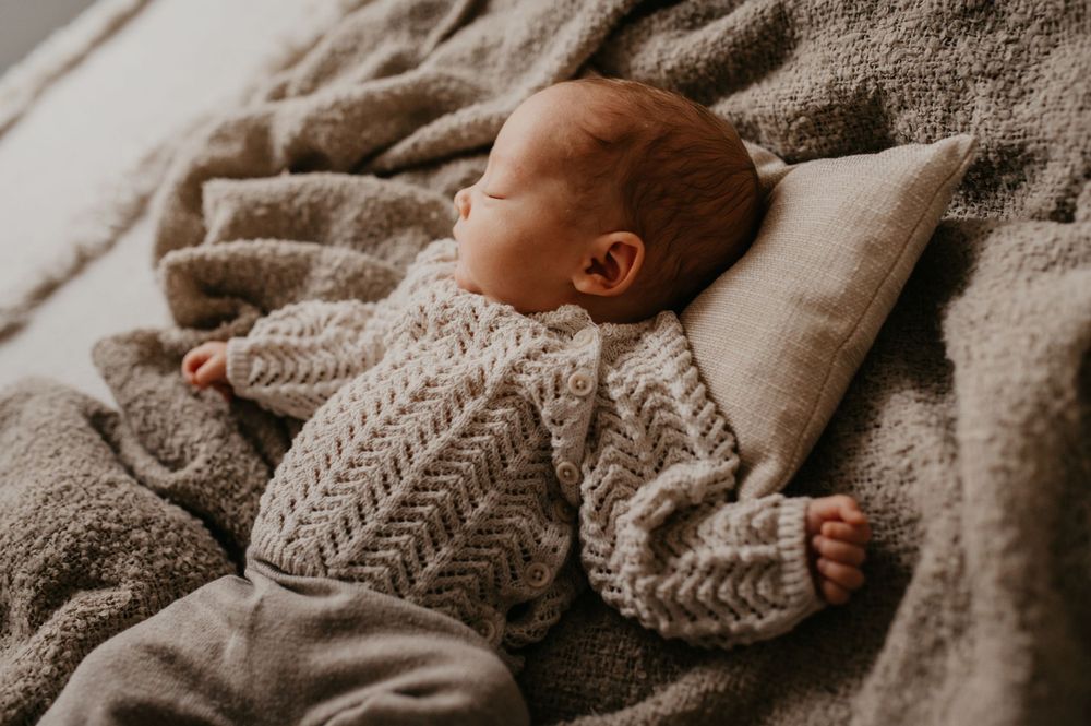 Baby sleeping peacefully on gray bedding wearing cozy knit sweater in natural morning light.