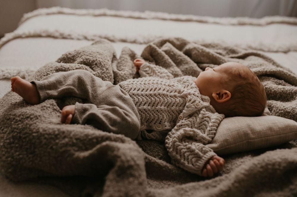 An infant peacefully sleeps on soft gray blankets wearing a cozy knitted sweater.