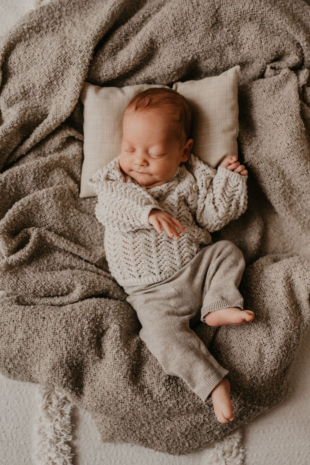 Sleeping baby wearing knit sweater and gray pants lies peacefully on soft brown blanket with small white pillow.