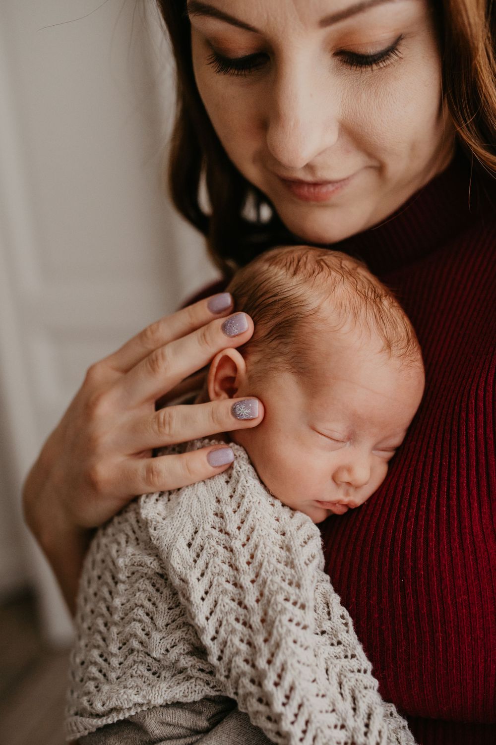 A tender moment shows a parent in a burgundy sweater cradling a sleeping newborn wearing a soft knit sweater.