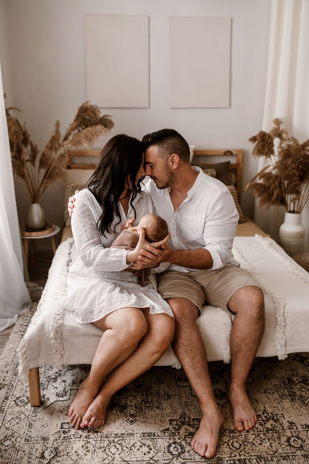 A couple sits on a bed with pampas grass decor, wearing white and beige outfits in a neutral-toned boho bedroom setting.