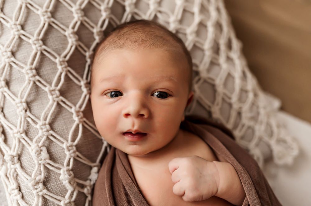 Newborn baby wrapped in brown fabric lying on textured white blanket during professional photo session.