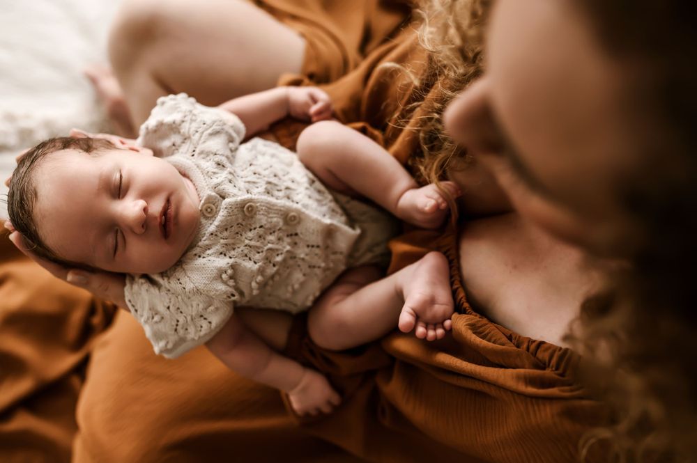 Sleeping baby in white lace dress lying on warm brown textured fabric in soft natural lighting.
