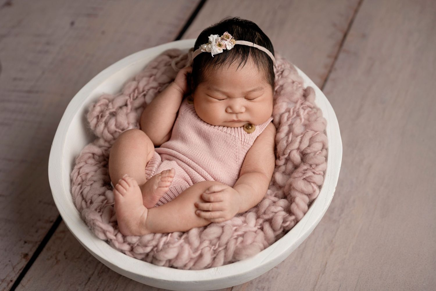 Sleeping newborn baby wrapped in soft pink fabric lying in white bowl against rustic wooden background.