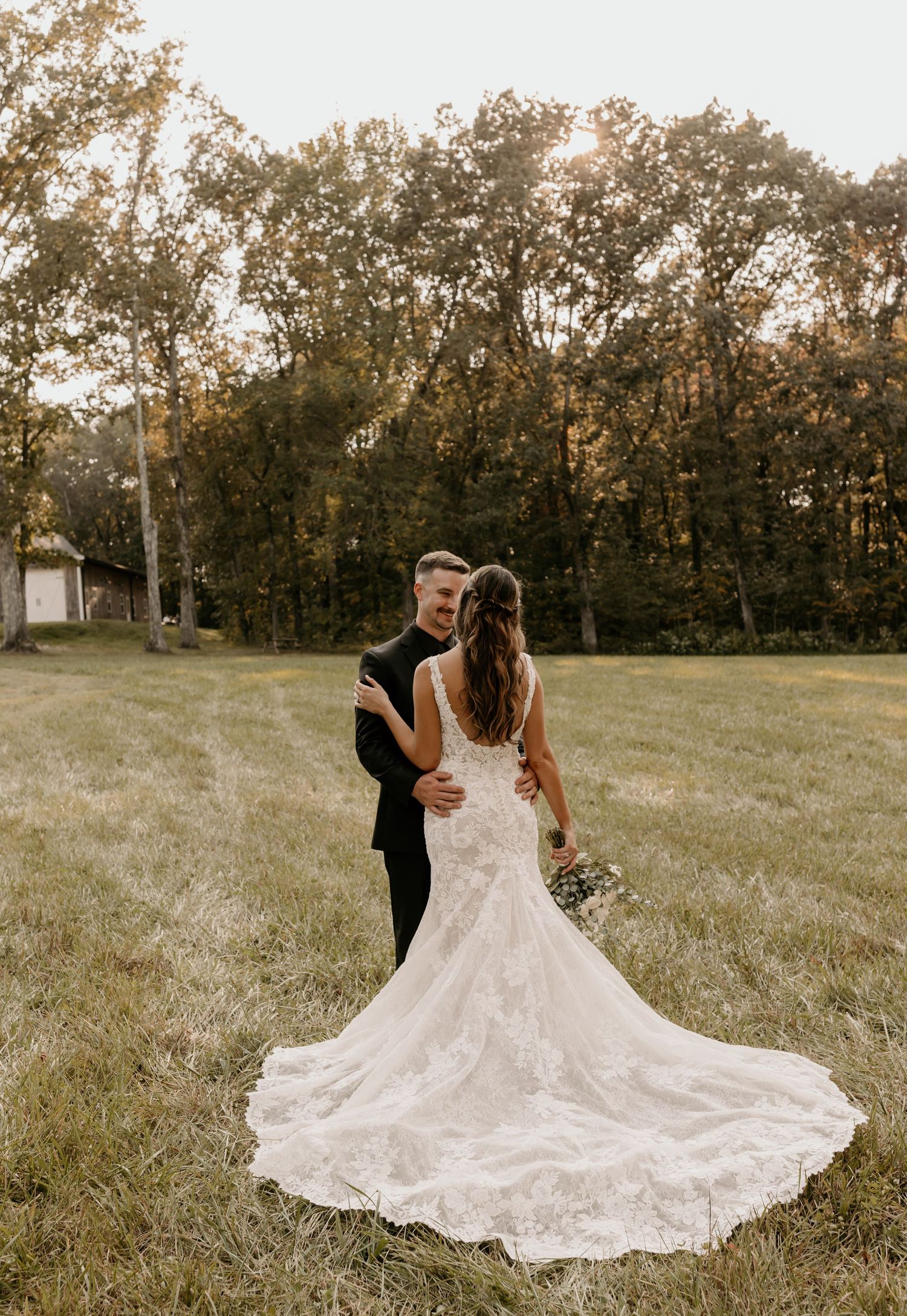 A stunning wedding dress with lace train trails behind couple in autumn field.