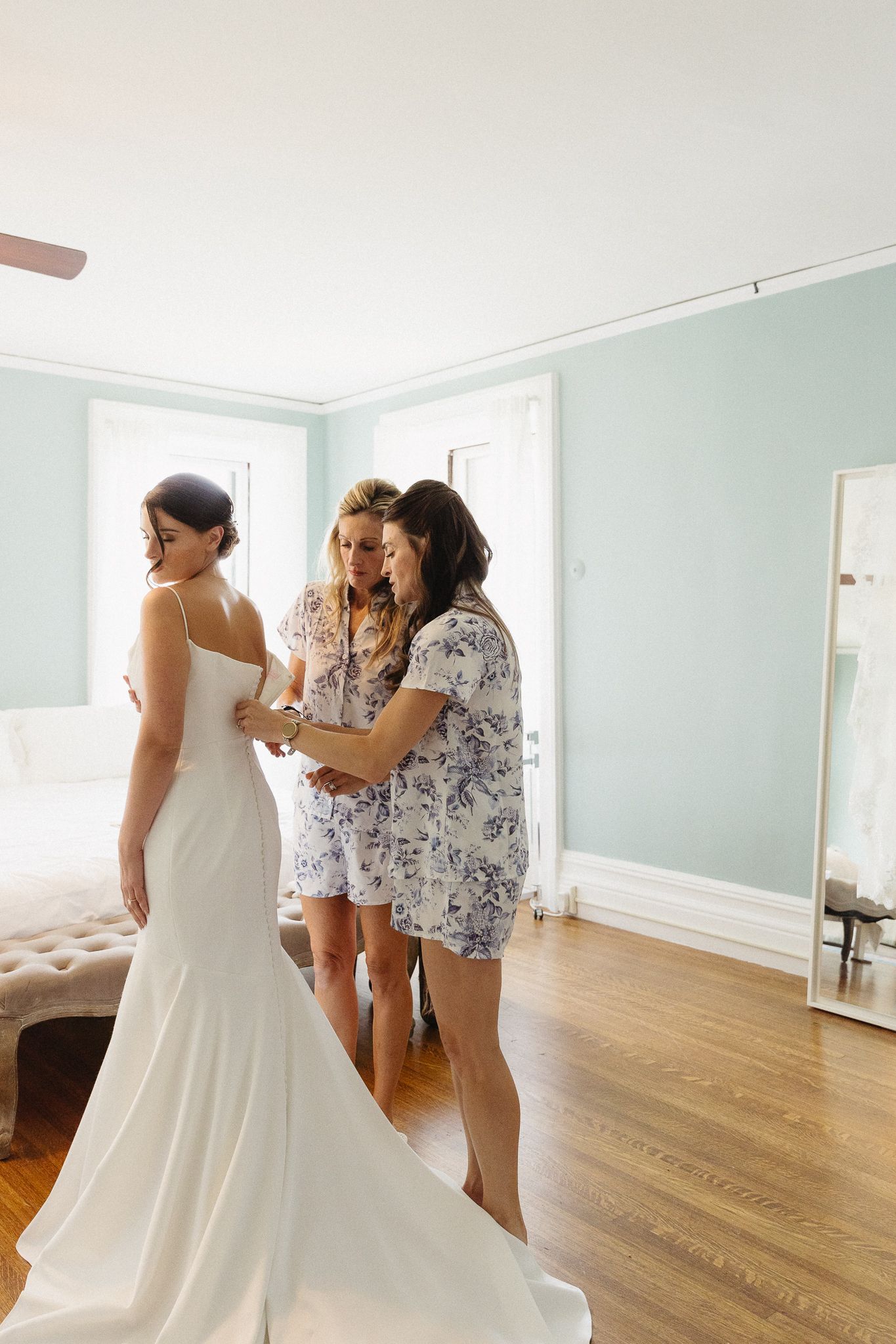 Getting ready scene with someone in a fitted white wedding gown being helped by others.