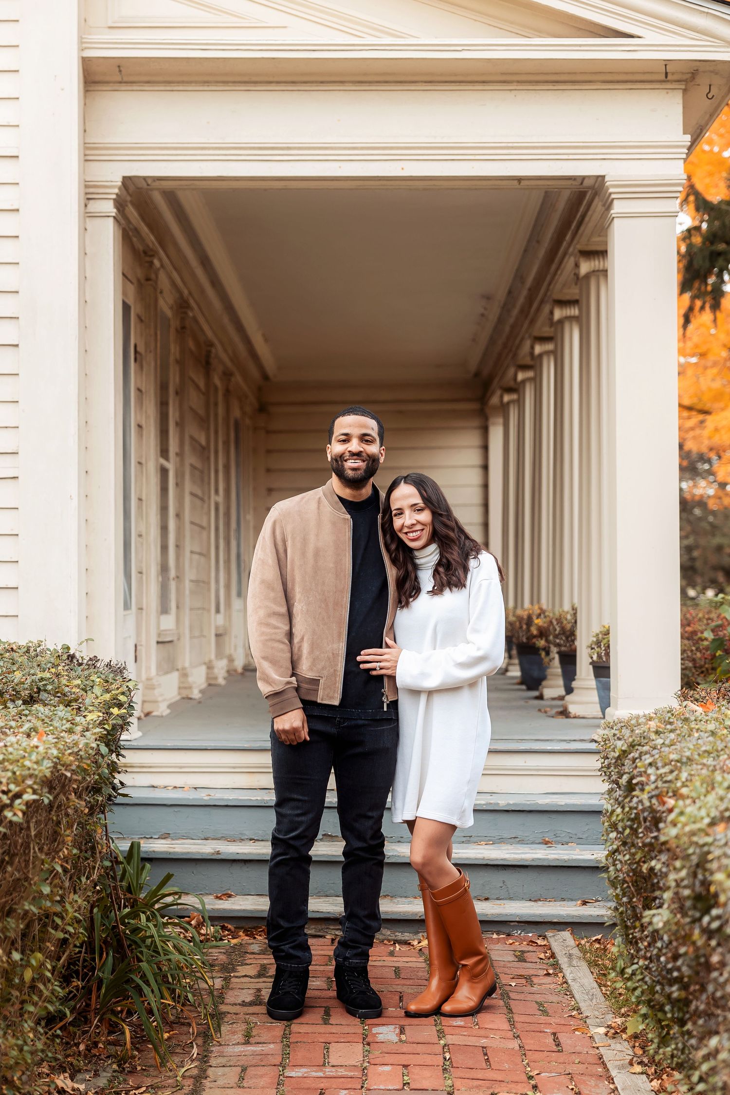 A couple poses together on a columned porch during fall season. Michigan Detroit Novi Northville Portrait Photographer