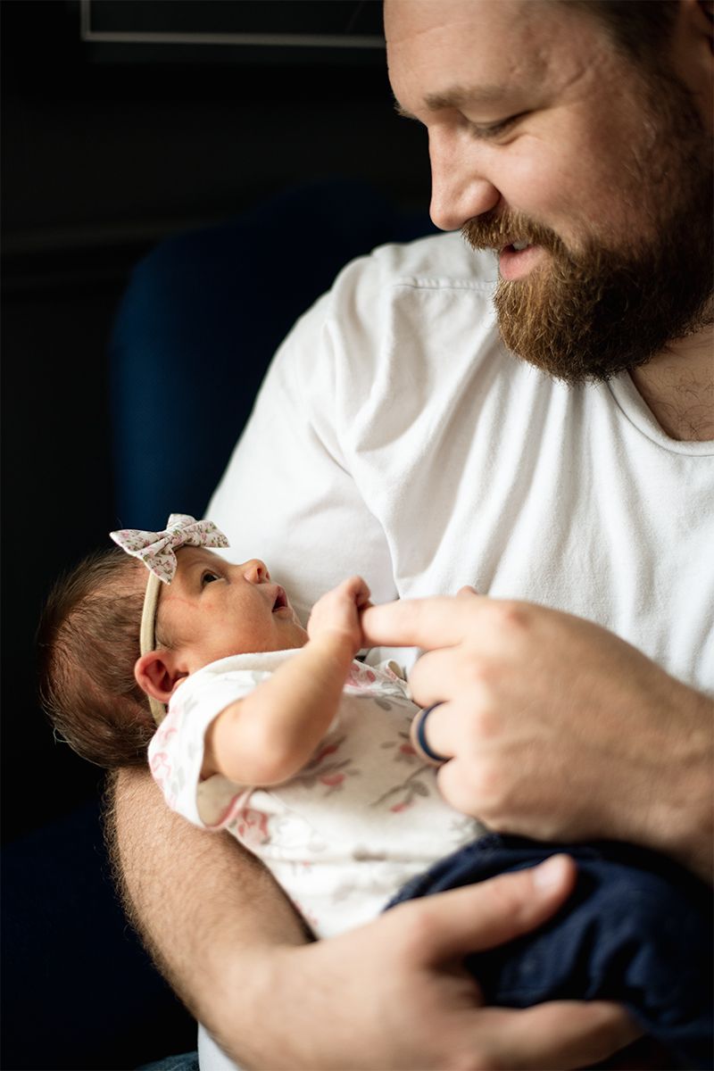 Bearded parent in white shirt lovingly holding and looking down at tiny newborn baby wearing bow.
