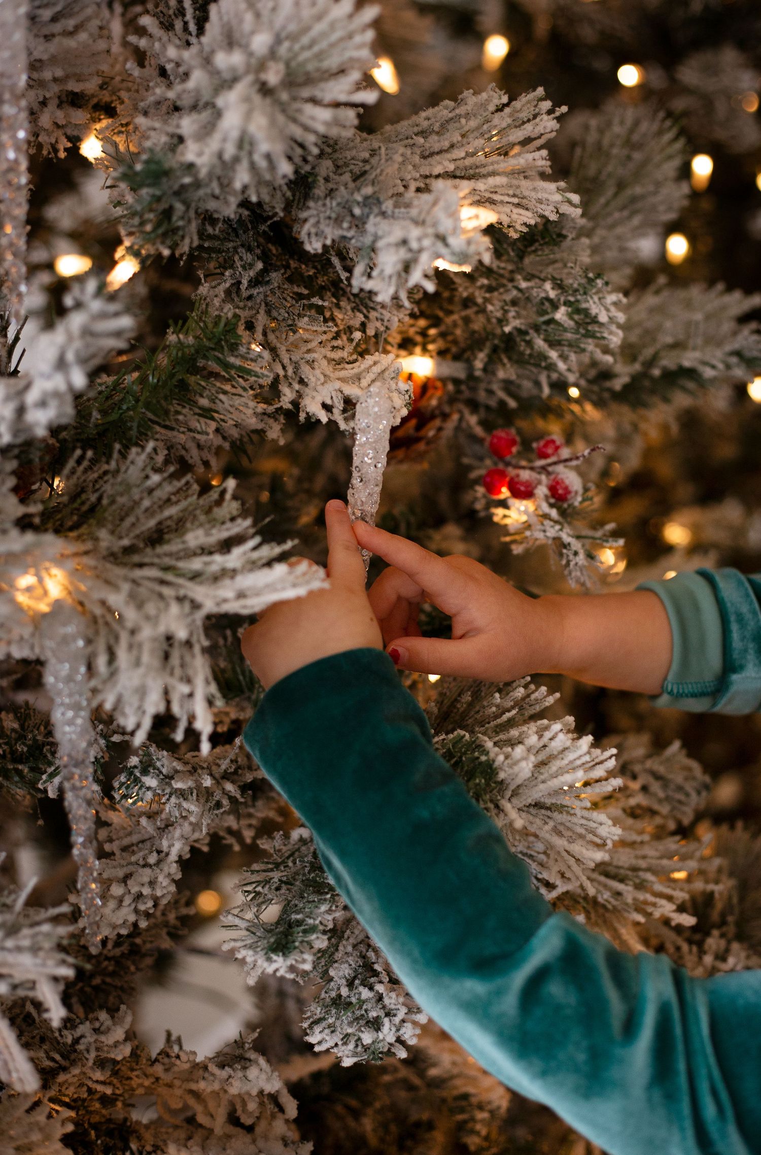 Hands in green sleeves decorate a snowy flocked Christmas tree with twinkling lights and winter ornaments.