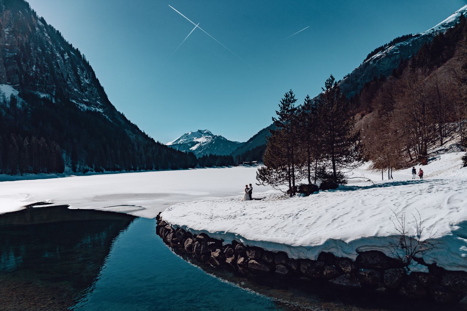 Snow-covered landscape with mountains and trees reflecting in partially frozen lake under blue sky in the French Alps.
