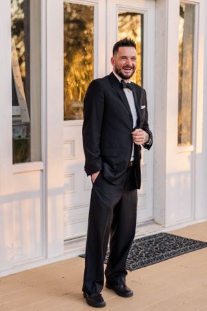 A groom in classic black tuxedo stands smiling on a porch near white doors.