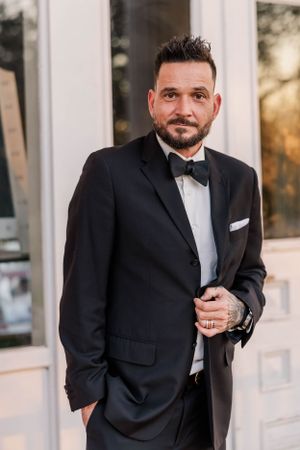A groom in black bow tie and tuxedo poses against white doors at sunset.