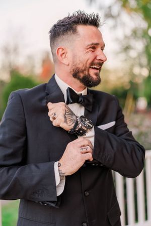 A groom in a black tuxedo smiles while adjusting his bowtie outdoors.