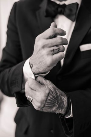 Close-up of tattooed hands adjusting cufflinks on a black tuxedo sleeve.
