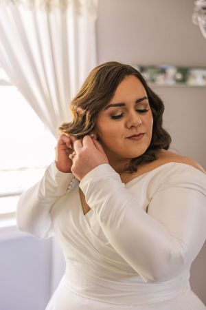 Bride adjusts earrings while getting ready in elegant white dress.