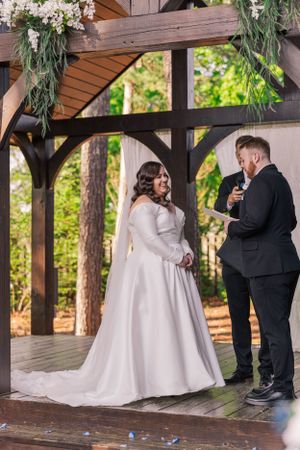 Wedding ceremony taking place under wooden pavilion surrounded by pine trees.