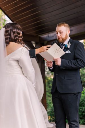 Couple sharing intimate moment during wedding ceremony while reading vows under wooden pergola.