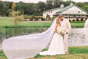 A wedding couple poses by a lake with a flowing cathedral veil and white venue building in the background.