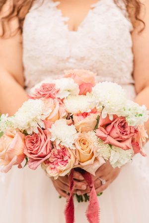 A delicate bridal bouquet features coral roses and white carnations with pink ribbon accents.