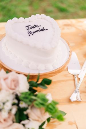 A heart-shaped white wedding cake with 'Just Married' text sits on a wooden table with floral decorations.
