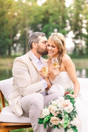 A wedding couple shares champagne while sitting outdoors by a lake with floral arrangements.