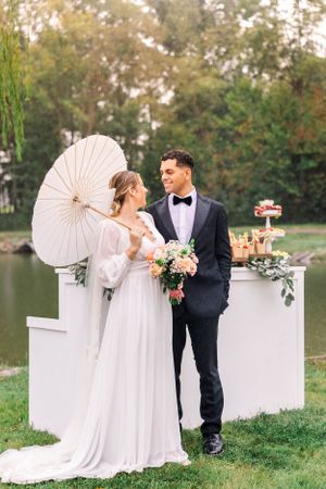 Elegant wedding setup by lake with white parasol and floral-decorated dessert bar.